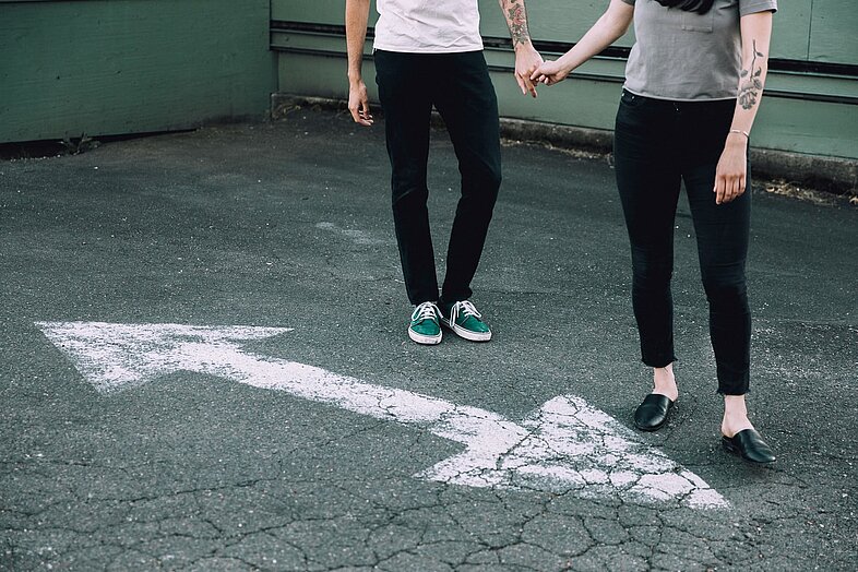 Symbolbild Paar: Zwei Personen halten Händchen über einem Richtungspfeil auf Asphalt