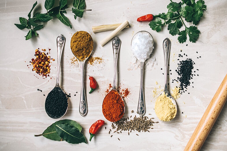 Verschiedene ayurvedische Gewürze auf Löffeln – Kurkuma, Kreuzkümmel, Senfsamen, Pfeffer und mehr – auf hellem Untergrund