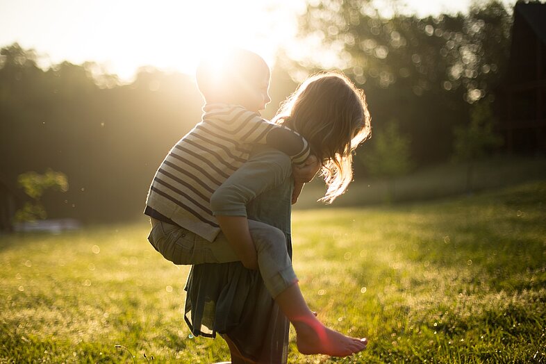 Erwachsener trägt ein Kind huckepack auf einer sonnigen Wiese als Symbolbild zur Ayurveda-Familienküche