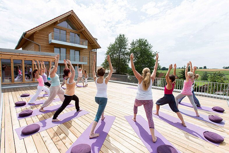 Yoga-Gruppe auf der Holzterrasse vor dem modernen Holzhaus des Rosenberg Ayurveda Zentrums in der Krieger-Pose mit erhobenen Armen